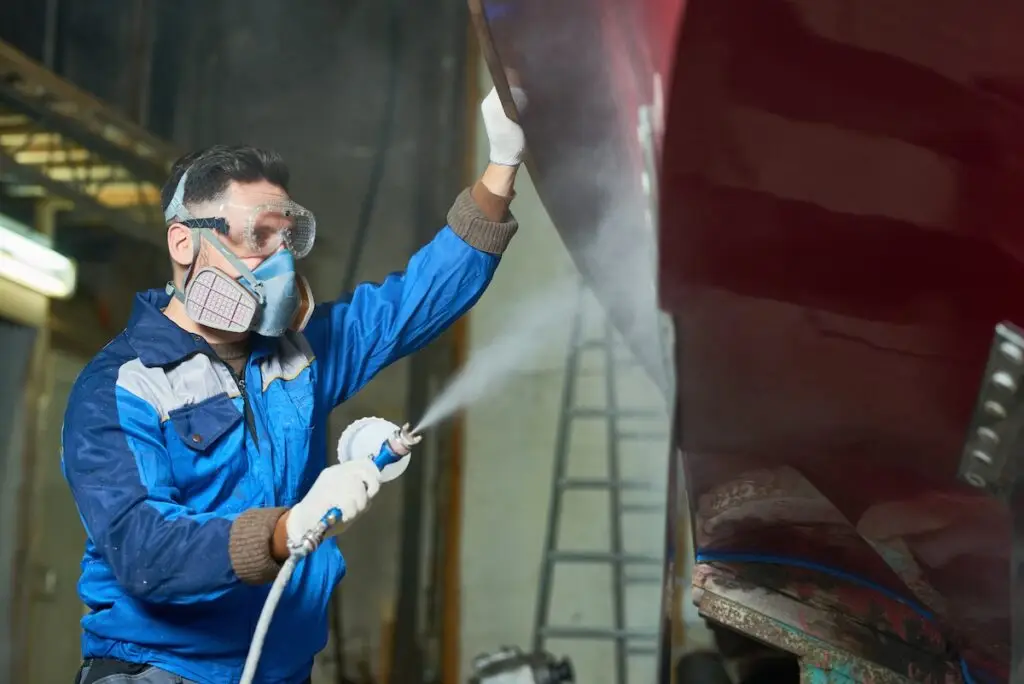 Side view portrait of worker wearing protective mask spray painting boat in yacht workshop